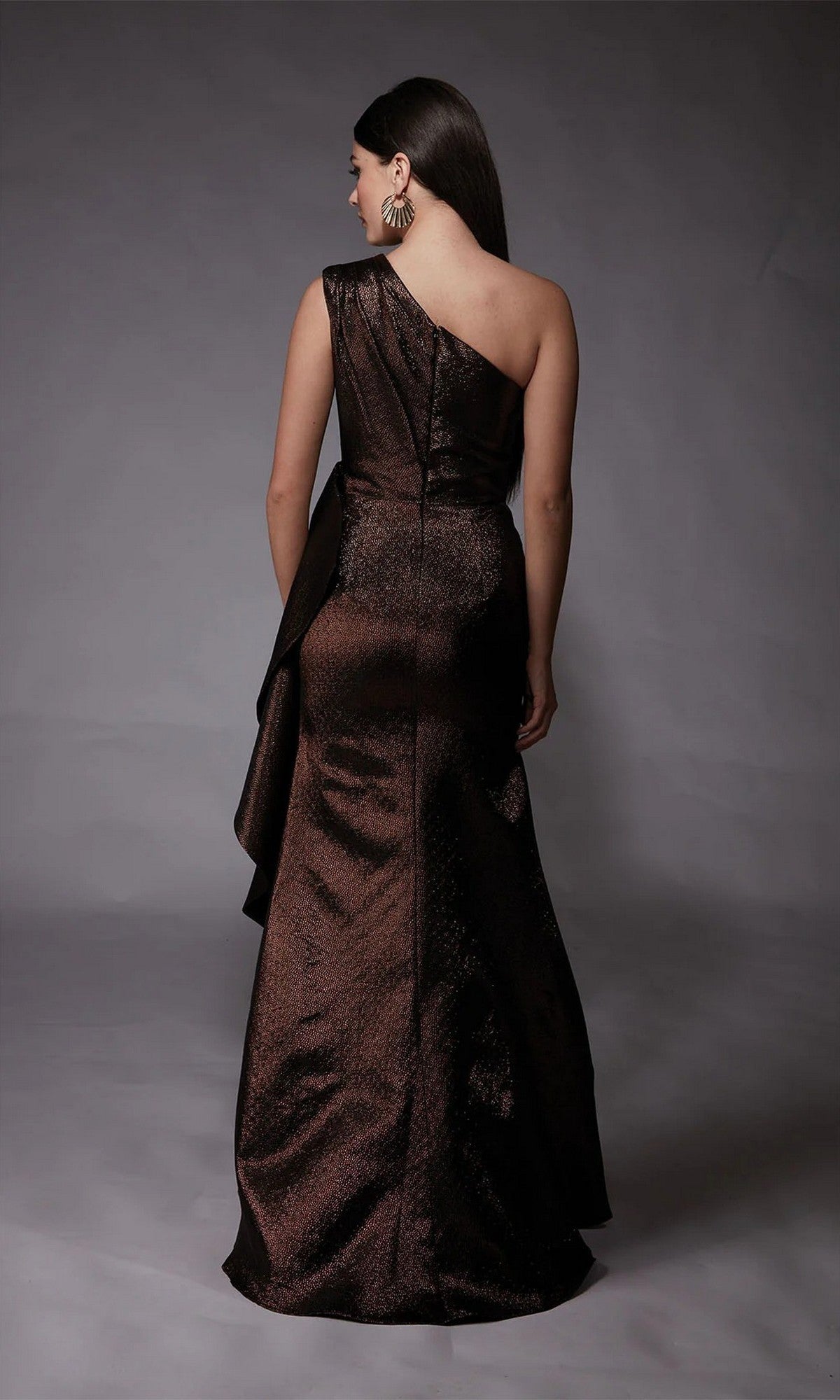 A woman stands facing away, wearing the shiny dark brown Alyce 27751 long formal jacquard one-shoulder dress with a floor-length skirt, against a plain gray background.